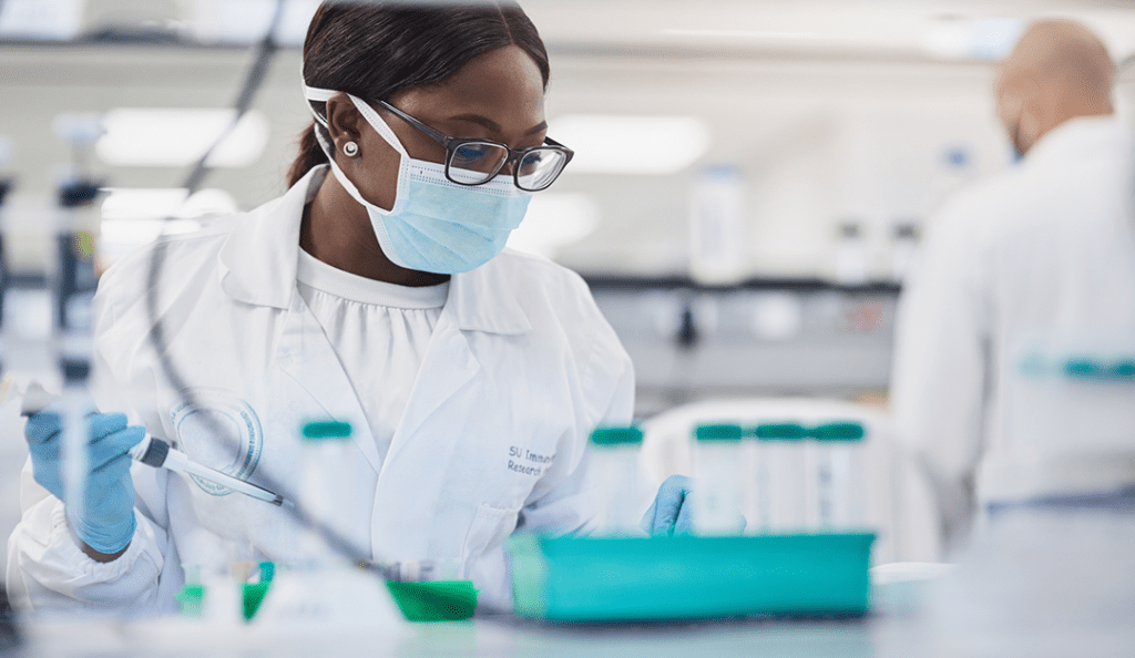 a woman researcher works in a lab wearing a mask and lab coat