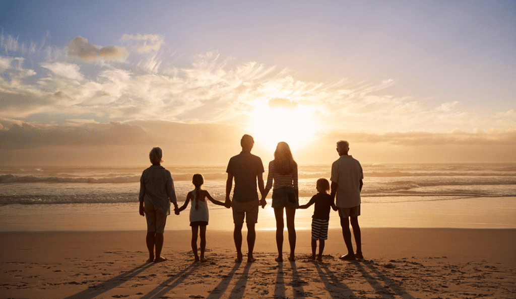 Several generations of a family stand on a beach facing away looking at sunset