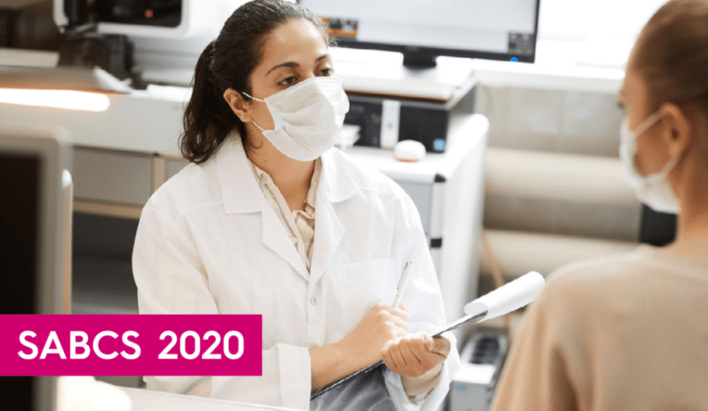Doctor wearing a lab coat and mask holding a clip board speaks to a patient