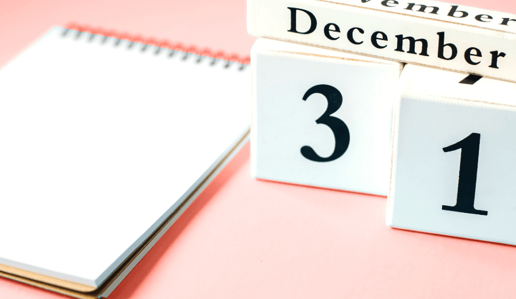 a wood block calendar shows the date december 31 next to a blank notebook and against a blush pink table
