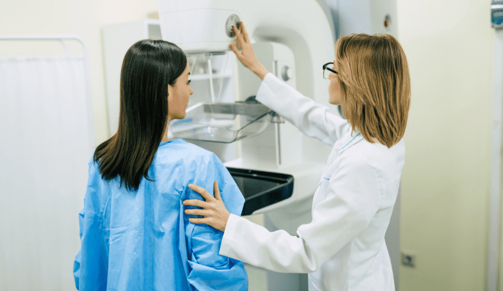 a woman in a blue medical gown prepares to get a mammogram as a tech assists wearing a white lab coat