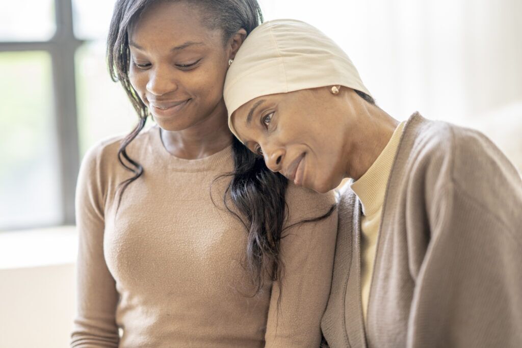 A young woman of African decent, sits with a friend as she battles through Cancer treatments. She is sitting in close as she allows her friend to rest her head on her shoulder. The patient is dressed comfortably and wearing a headscarf to keep her warm. The two have neutral expressions on their faces.