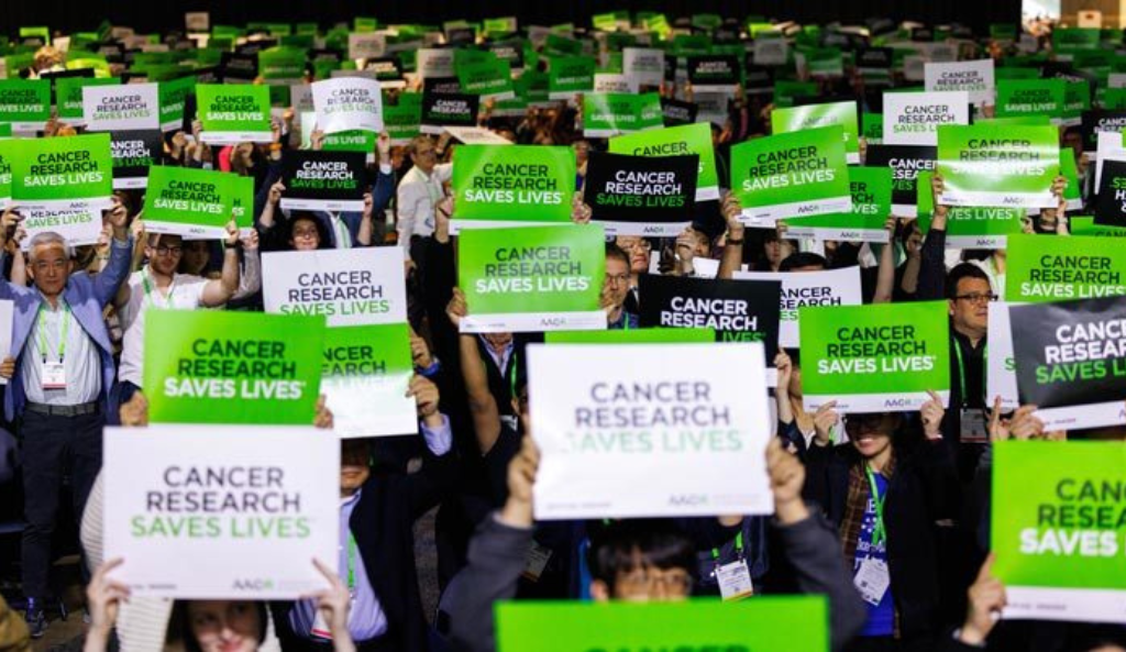 AACR 2025 meeting attendees hold up signs that read CANCER RESEARCH SAVES LIVES