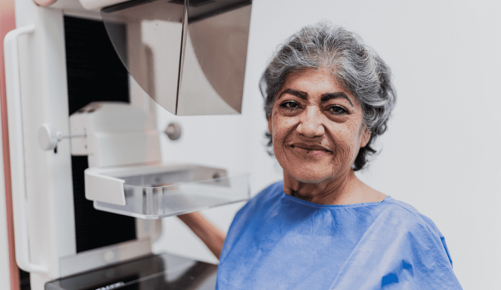 a woman prepares for her mammogram, she stands in front of the machine and is smiling at the camera wearing a blue gown
